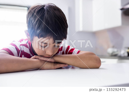 Boy leaning head on folded arms on white kitchen table at home near bright window cabinet 133292473
