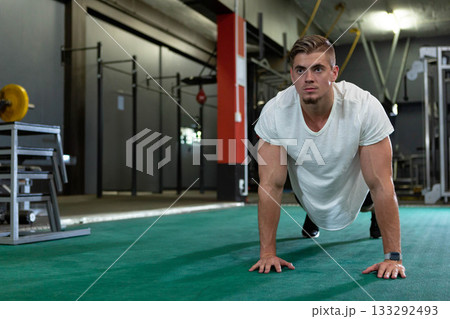 Man doing push-up on padded gym floor wearing sportswear and smartwatch with barbell rack behind Man doing push-up on padded gym floor wearing sportswear and smartwatch with barbell rack behind 133292493