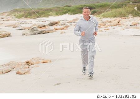 Adult man running along sandy shoreline wearing light grey hoodie grey sweatpants and running shoes 133292502