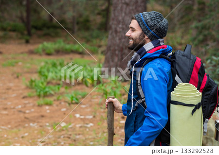 Man hiking in pine forest with backpack, holding walking stick and wearing knit cap, copy space Man hiking in pine forest with backpack, holding walking stick and wearing knit cap, copy space 133292528