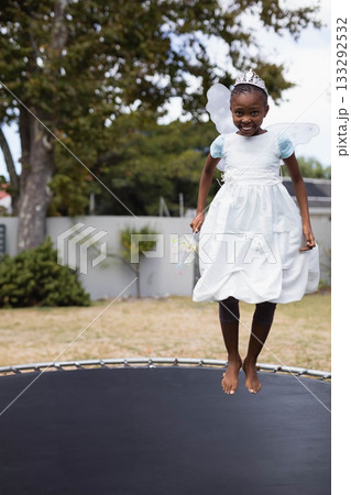 African American girl bouncing on yard trampoline wearing tiara and fairy wings holding ribbon wand 133292532