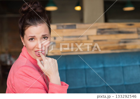 Teenage female wearing blazer biting finger on bench by slat wall in cafe with pendant lights Teenage female wearing blazer biting finger on bench by slat wall in cafe with pendant lights 133292546