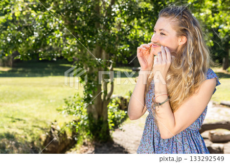 Woman standing in park holding grass blade under nose wearing bracelets, looking right, copy space Woman standing in park holding grass blade under nose wearing bracelets, looking right, copy space 133292549