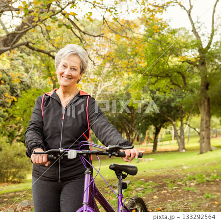 Senior woman standing under leafy tree in park holding purple bicycle handlebars sporting jacket 133292564