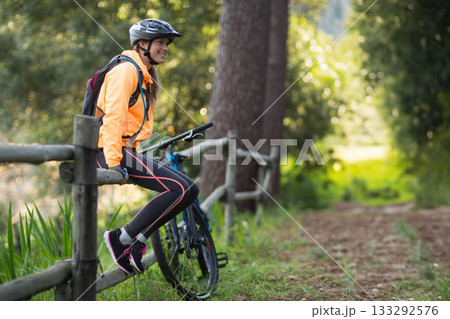 Female cyclist wearing helmet sitting on wooden fence at forest trail holding bike, copy space 133292576