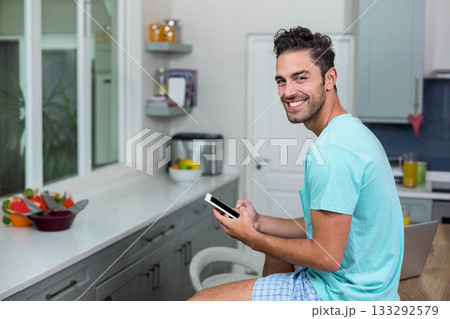 Man sitting on stool at kitchen counter holding smartphone and laptop with fruit bowl, copy space 133292579