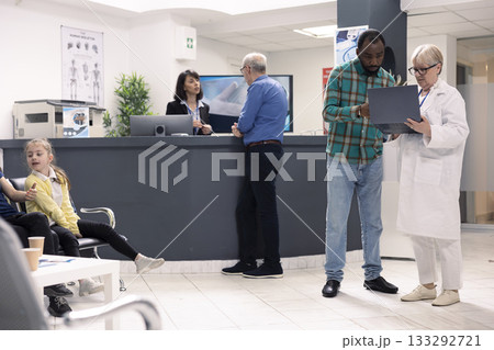 Elderly female physician in lab coat consults with male patient in hospital waiting room. Senior doctor with laptop discusses recovery plan with black man as receptionist assists clinic visitors. Elderly female physician in lab coat consults with male patient in hospital waiting room. Senior doctor with laptop discusses recovery plan with black man as receptionist assists clinic visitors. 133292721