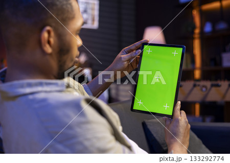 Black freelancer uses green screen mockup on modern tablet to work in the living room, sitting at home with the gadget and solving tasks. Relaxed casual young adult in his trendy space. Black freelancer uses green screen mockup on modern tablet to work in the living room, sitting at home with the gadget and solving tasks. Relaxed casual young adult in his trendy space. 133292774