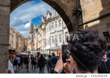 Prague, Czech Republic, August 9, 2023. A young man takes photos with his cell phone from Charles Bridge toward the Mala Strana neighborhood. The colorful facades stand out. Crowded with people. 133293039