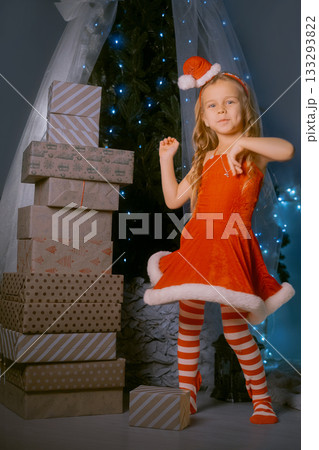 Young girl in a red Santa dress dancing joyfully near a mountain of Christmas presents by a beautifully decorated tree. 133293822
