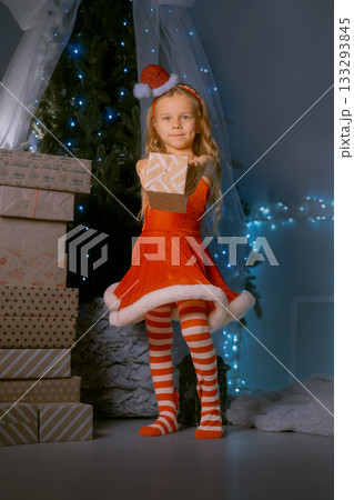 Christmas magic moment with a woman in red Santa dress standing by a brightly lit tree holding a holiday present. 133293845