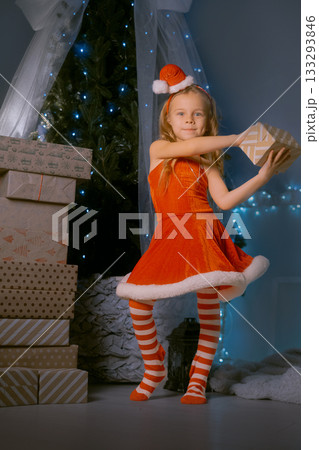 Festive holiday scene with a smiling girl in a Santa outfit presenting a wrapped gift near a Christmas tree 133293846