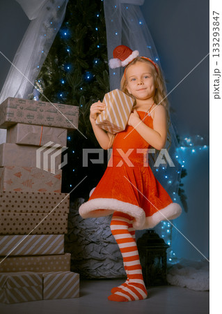 Young woman in a red Santa dress holding a Christmas present by a beautifully decorated tree with sparkling lights. 133293847