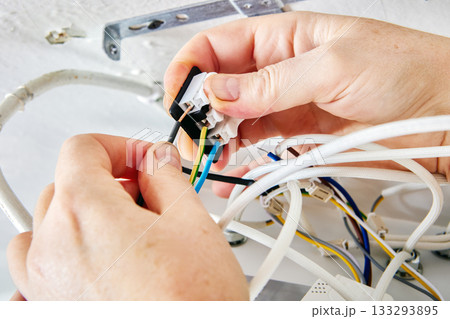 Mounting process involves technician joining electrical wires to terminal block of LED light fitting on ceiling surface in countryside home, device partially open. 133293895