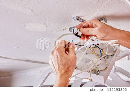 Wiring is being connected by electrician during installation of new LED chandelier on white ceiling in rural house, wires and fixture visible in frame. 133293896