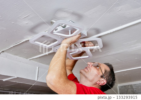 Lighting installation work includes electrician fitting LED chandelier to ceiling in rural house during setup of new lighting fixture. Lighting installation work includes electrician fitting LED chandelier to ceiling in rural house during setup of new lighting fixture. 133293915