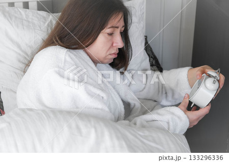 A woman, wrapped in a soft robe, with a wary expression on her face, looking at the alarm clock, among light pillows and a calm interior, anticipating the silence of the early morning. Copy space A woman, wrapped in a soft robe, with a wary expression on her face, looking at the alarm clock, among light pillows and a calm interior, anticipating the silence of the early morning. Copy space 133296336