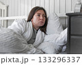 A woman, wrapped in a soft robe, with a wary expression on her face, looking at the alarm clock, among light pillows and a calm interior, anticipating the silence of the early morning. Copy space 133296337