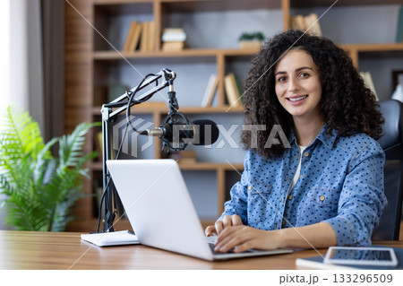 Young woman with curly hair hosting a podcast in her modern home office, smiling while using a laptop and professional microphone for remote work and online communication 133296509