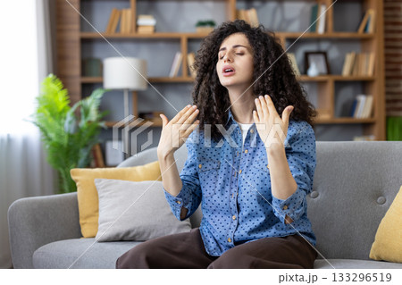 Woman feeling overheated and uncomfortable during a heatwave, fanning herself with hands while sitting on a sofa in a modern apartment, showing distress from high summer temperatures 133296519