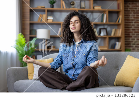 Woman sitting in lotus pose on a living room sofa with eyes closed and hands in mudra, finding peace and reducing stress through mindfulness and relaxation at home 133296523