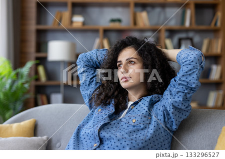Young woman with curly hair reclining on a gray couch in a modern living room, hands behind head, looking away thoughtfully, relaxed and contemplative during a peaceful break 133296527