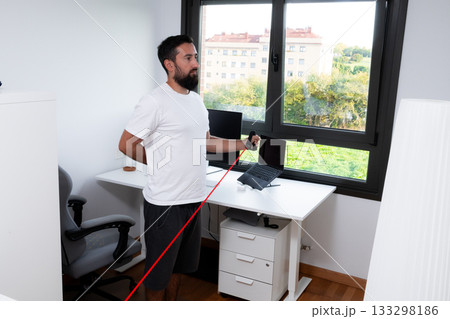 Young caucasian male exercising with resistance band in home office setting Young caucasian male exercising with resistance band in home office setting 133298186