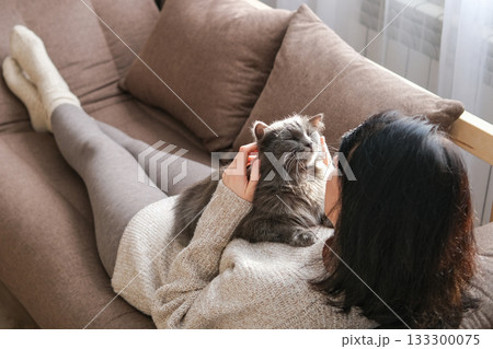 Woman is lying on brown sofa cuddling and stroking fluffy gray cat resting on her chest. Pet companionship, relaxation, cozy home life and comfort with animals Woman is lying on brown sofa cuddling and stroking fluffy gray cat resting on her chest. Pet companionship, relaxation, cozy home life and comfort with animals 133300075