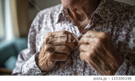 Senior woman with trembling hands struggling to button shirt, showing symptoms of aging, arthritis or neurological condition in soft indoor light 133300196