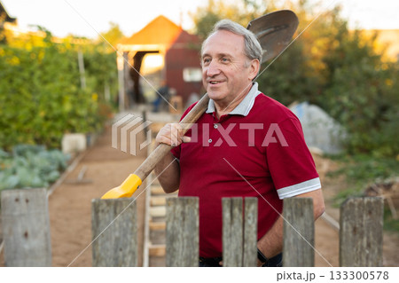 Mature man standing relaxed near fence with shovel in backyard smiling 133300578