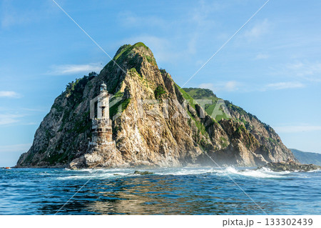 Aniva former japanese lighthouse photographed from sea. Abandoned tower stands at base of steep rocky cliffs with visible erosion, strong waves and coastal spray in clear daylight, Sakhalin island 133302439