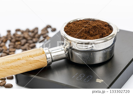 Portafilter with finely ground coffee on digital kitchen scale showing 18 grams weight beside scattered coffee beans on white Portafilter with finely ground coffee on digital kitchen scale showing 18 grams weight beside scattered coffee beans on white 133302720