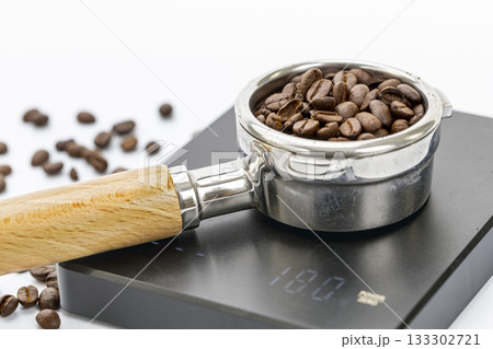 Close-up of roasted coffee beans in an espresso portafilter on a digital coffee scale with scattered beans on white background 133302721