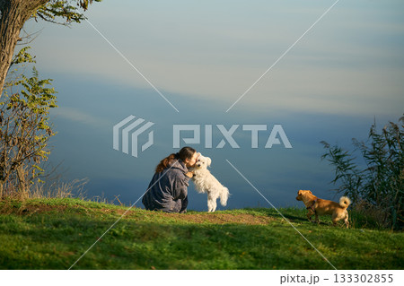Woman kiss and hug her cute white dog sitting on grass over scenic mountain background 133302855