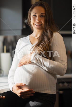 Smiling pregnant woman holding belly in a bright home kitchen Smiling pregnant woman holding belly in a bright home kitchen 133303249