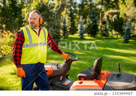 Male gardener standing near ride-on lawnmower on green grass 133304487