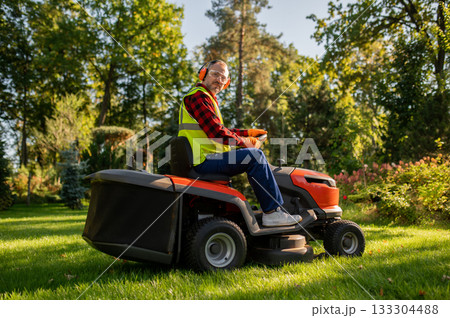 Worker in safety gear standing near lawn tractor in garden area 133304488