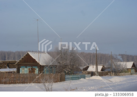 The photograph depicts old houses in a Siberian village, an old fence, and snowdrifts. The roofs of the houses are covered in a thick layer of snow, and a winter road leads through the village. 133306958