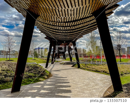 quiet urban park with wooden pathway, solitary individual strolling beneath shaded slatted roof 133307510