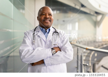 Smiling mature black doctor standing confidently with arms crossed in modern interior Smiling mature black doctor standing confidently with arms crossed in modern interior 133310455