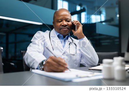 Mature African American doctor talking on cellphone while writing notes at desk Mature African American doctor talking on cellphone while writing notes at desk 133310524