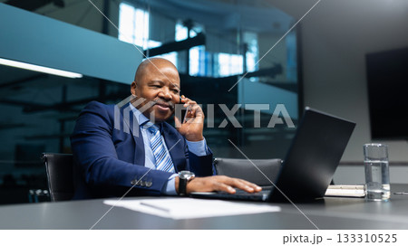Black businessman speaking on cellphone while working on laptop in modern office Black businessman speaking on cellphone while working on laptop in modern office 133310525