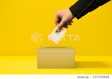 A person putting a voting card into a yellow ballot box 133310604