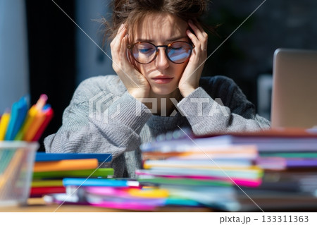 A student appears stressed while studying in a well-lit room filled with books and colorful school supplies. Her hands are on her head, showing her struggle to concentrate 133311363
