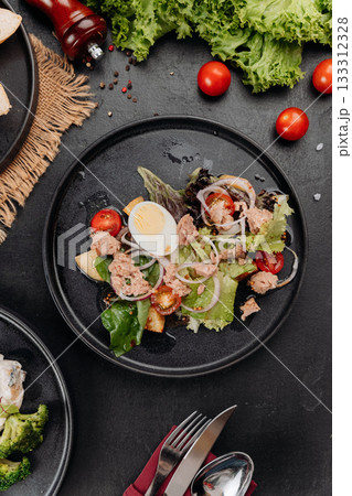 Tuna salad with egg, tomatoes and greens on black plate, restaurant lunch closeup 133312328