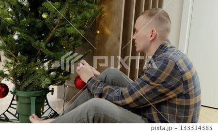 Young male decorating christmas tree with red baubles, sitting on floor, creating festive holiday scene in warmly lit home interior 133314385