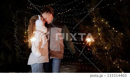 Couple holding sparkling fireworks, celebrating festive night amid illuminated winter forest with glowing holiday lights Couple holding sparkling fireworks, celebrating festive night amid illuminated winter forest with glowing holiday lights 133314403