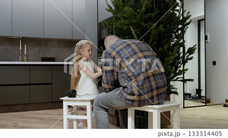 Father and daughter sitting on stools untangling christmas lights for the christmas tree in a modern apartment, preparing for the holiday season by decorating their home 133314405
