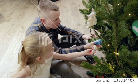 Family moment showing father helping young daughter hanging christmas ornaments, bonding while decorating festive tree together in cozy home living space Family moment showing father helping young daughter hanging christmas ornaments, bonding while decorating festive tree together in cozy home living space 133314407