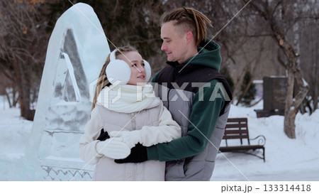 Loving couple wearing warm winter clothing embracing near intricate ice sculpture, snow covered park landscape creating romantic seasonal backdrop Loving couple wearing warm winter clothing embracing near intricate ice sculpture, snow covered park landscape creating romantic seasonal backdrop 133314418